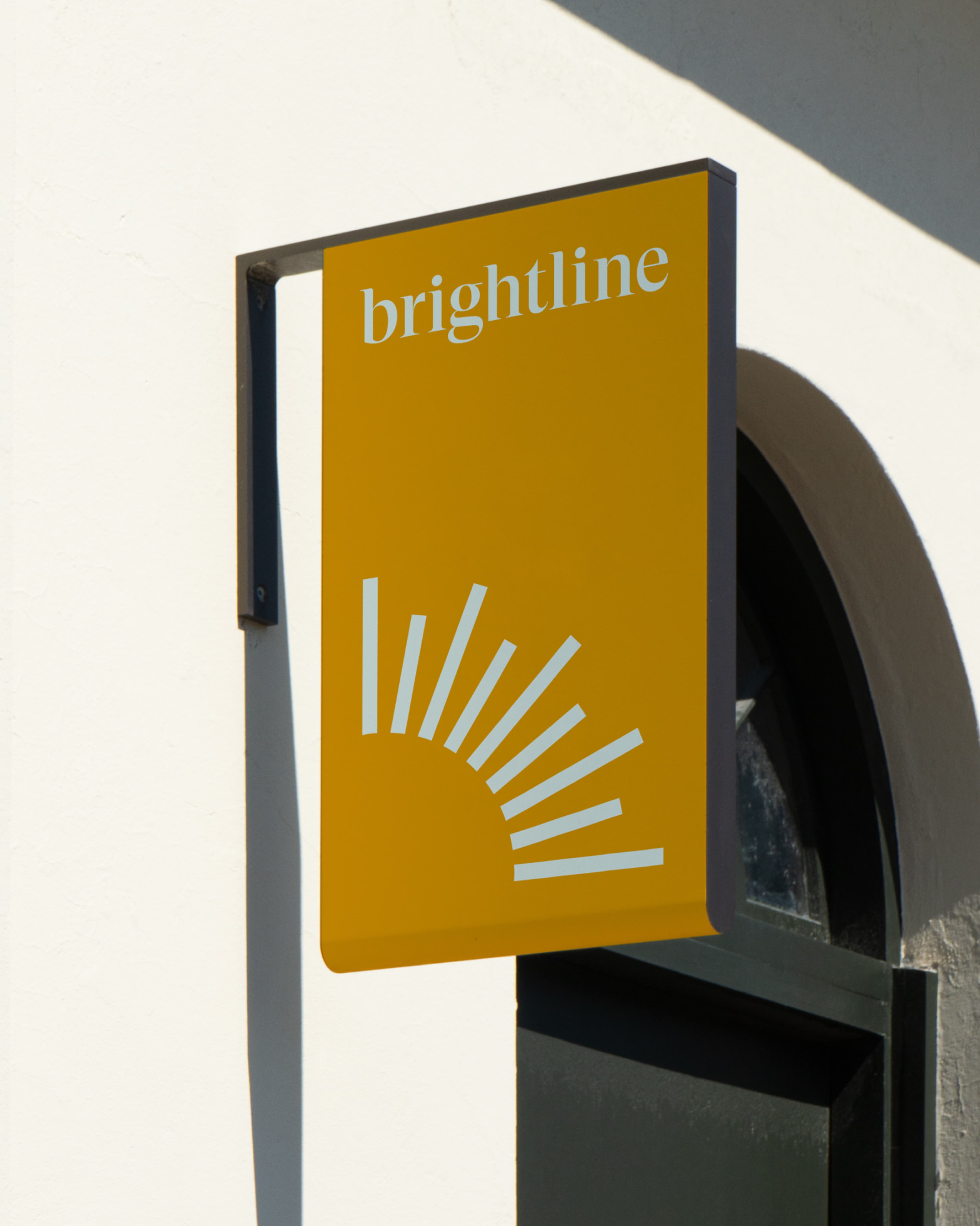 Yellow signboard with white text 'brightline' and a stylized sun, mounted on a white wall above an arched dark door.