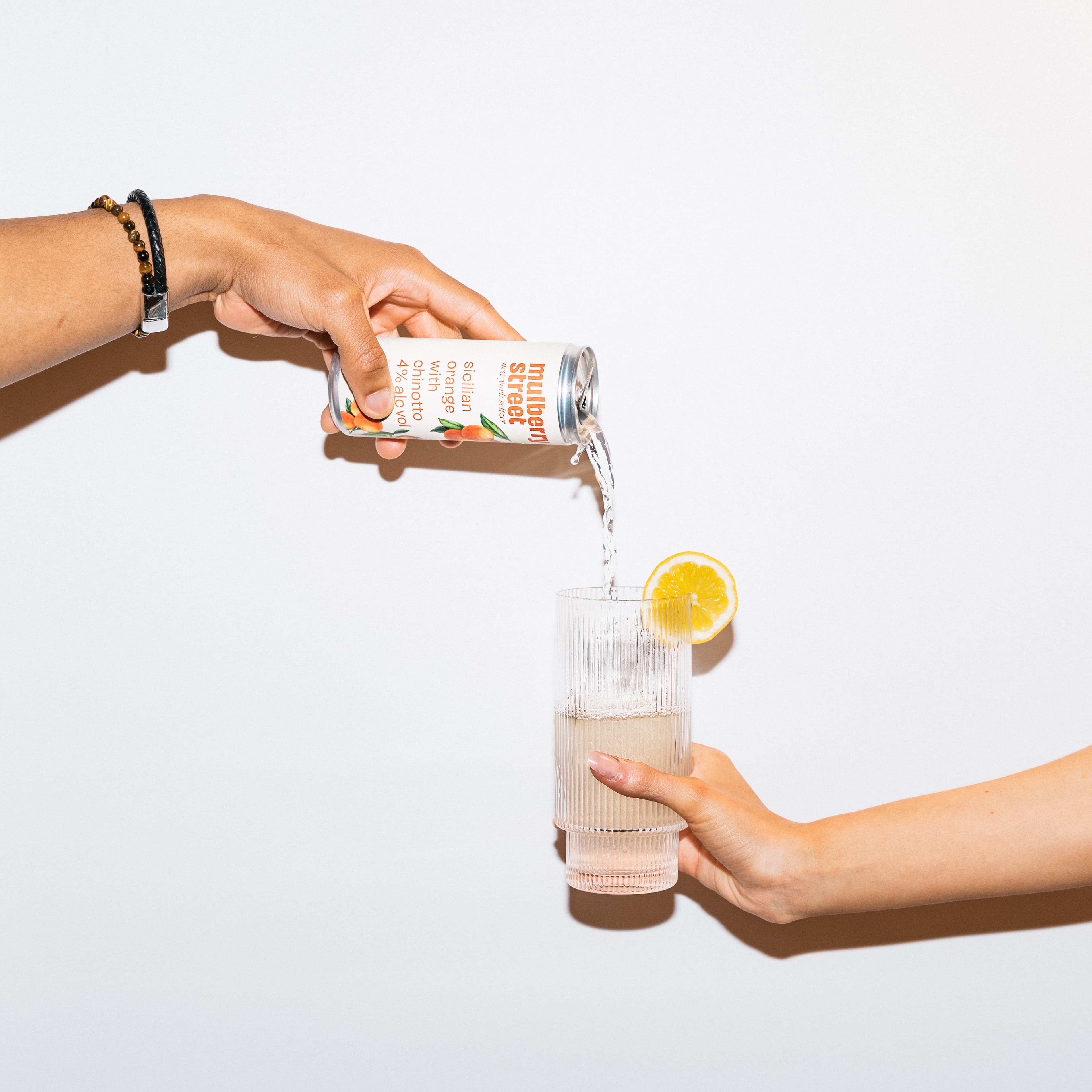 A hand pours a can of Mulberry Street Sicilian Orange with Chinotto hard seltzer (4% alcohol) into a ribbed glass garnished with a lemon slice, against a white background.