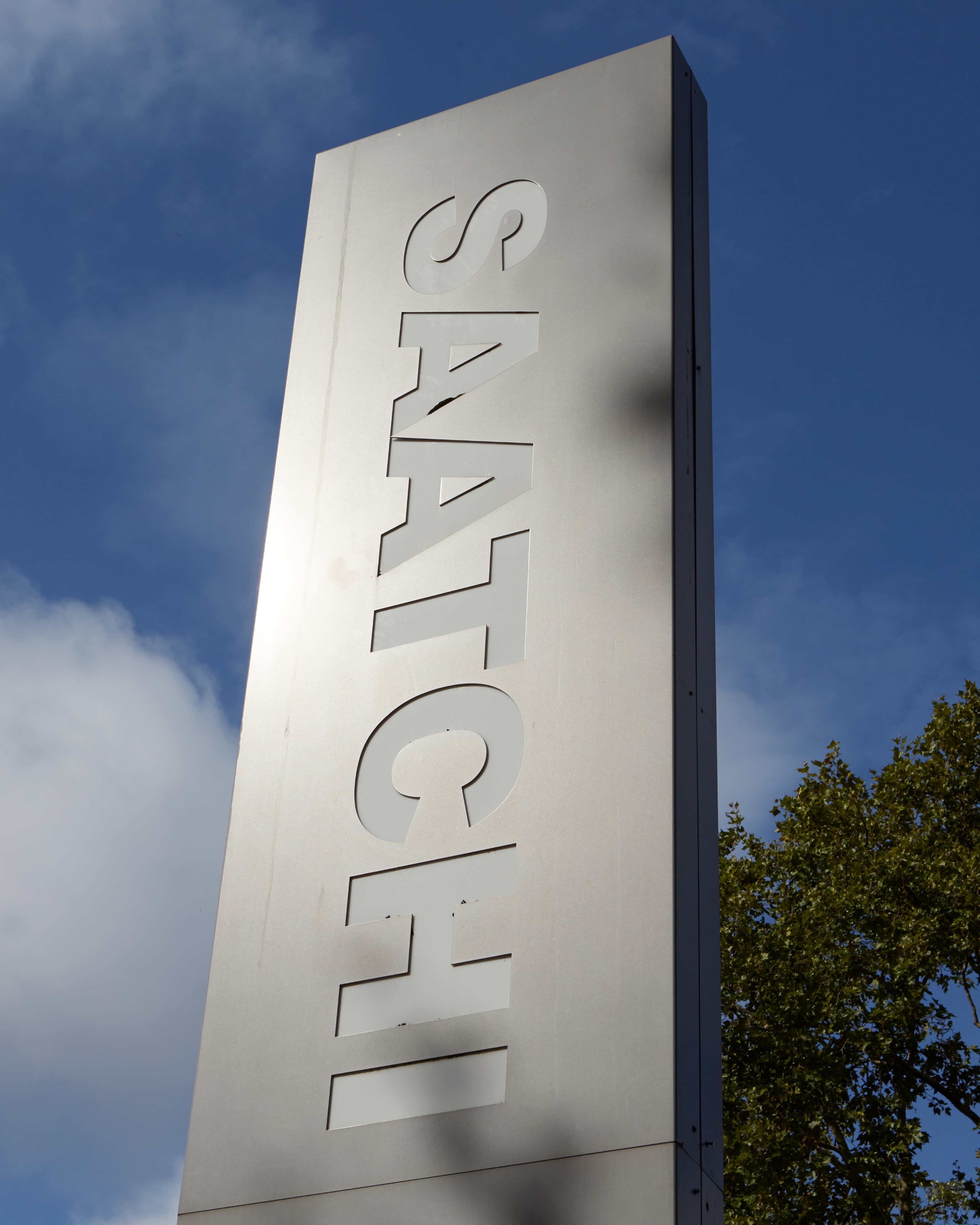 Tall metallic Saatchi sign against a blue sky with clouds and trees, reflecting sunlight.