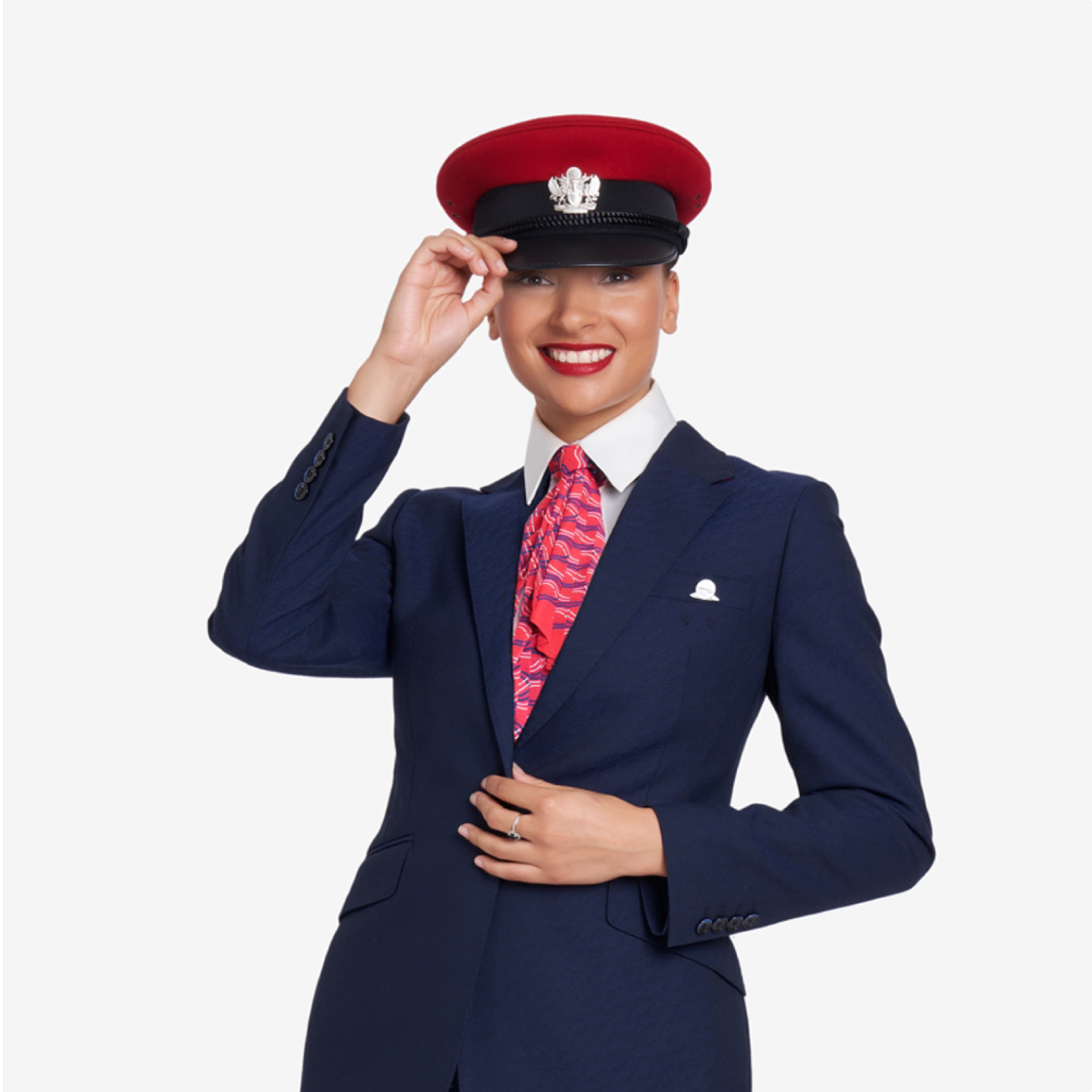 British Airways female cabin crew member in navy uniform, white shirt, red patterned necktie, and red-topped cap with BA emblem, smiling and tipping her cap.