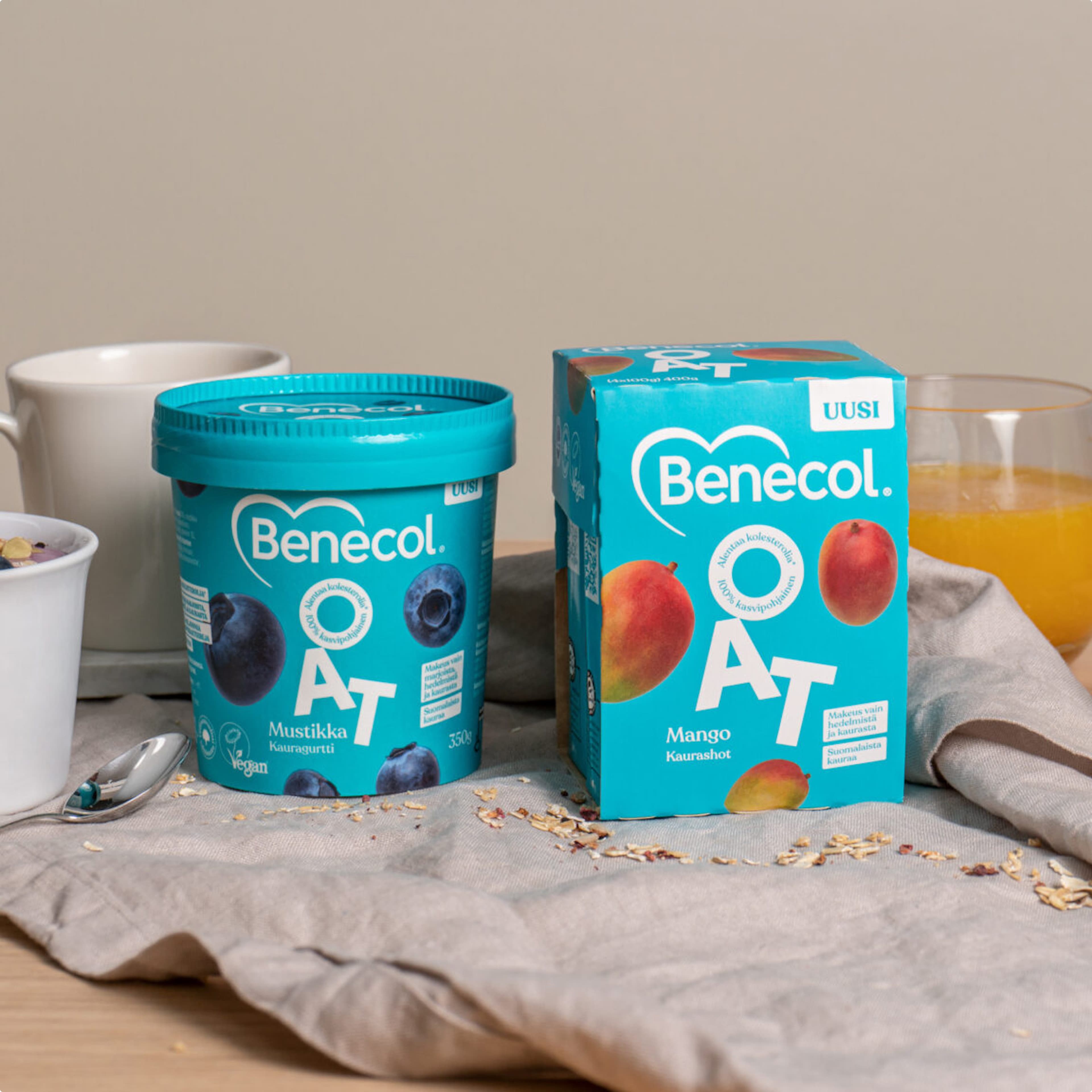 Two Benecol OAT products in teal packaging on a linen cloth: Mustikka (blueberry) Kauragurtti yogurt and Mango Kaurashot, surrounded by scattered oats, a bowl, mug, and glass of orange juice.