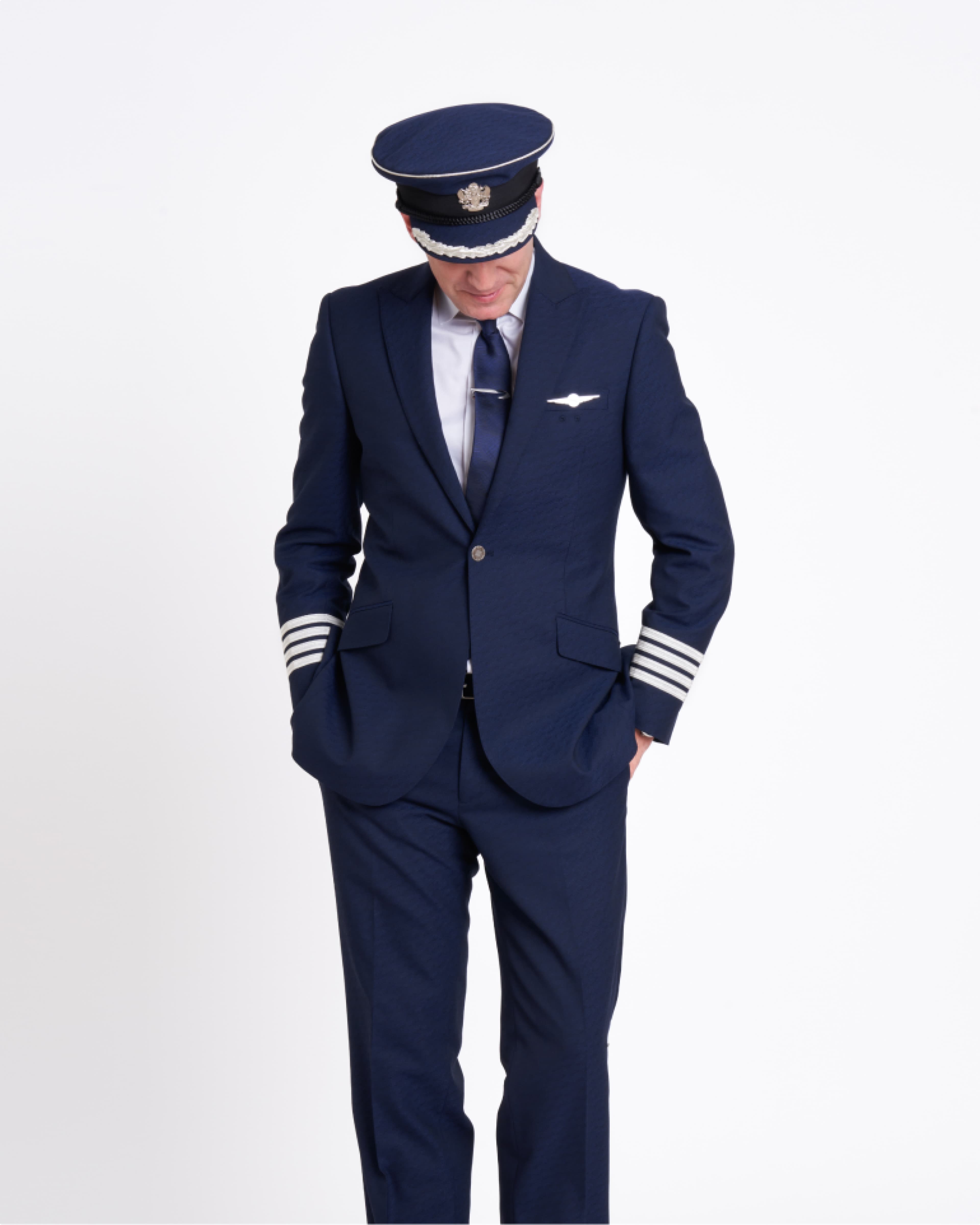 A pilot in a navy blue uniform with stripes on the sleeves and a matching hat, looking down, hands in pockets, against a plain background.