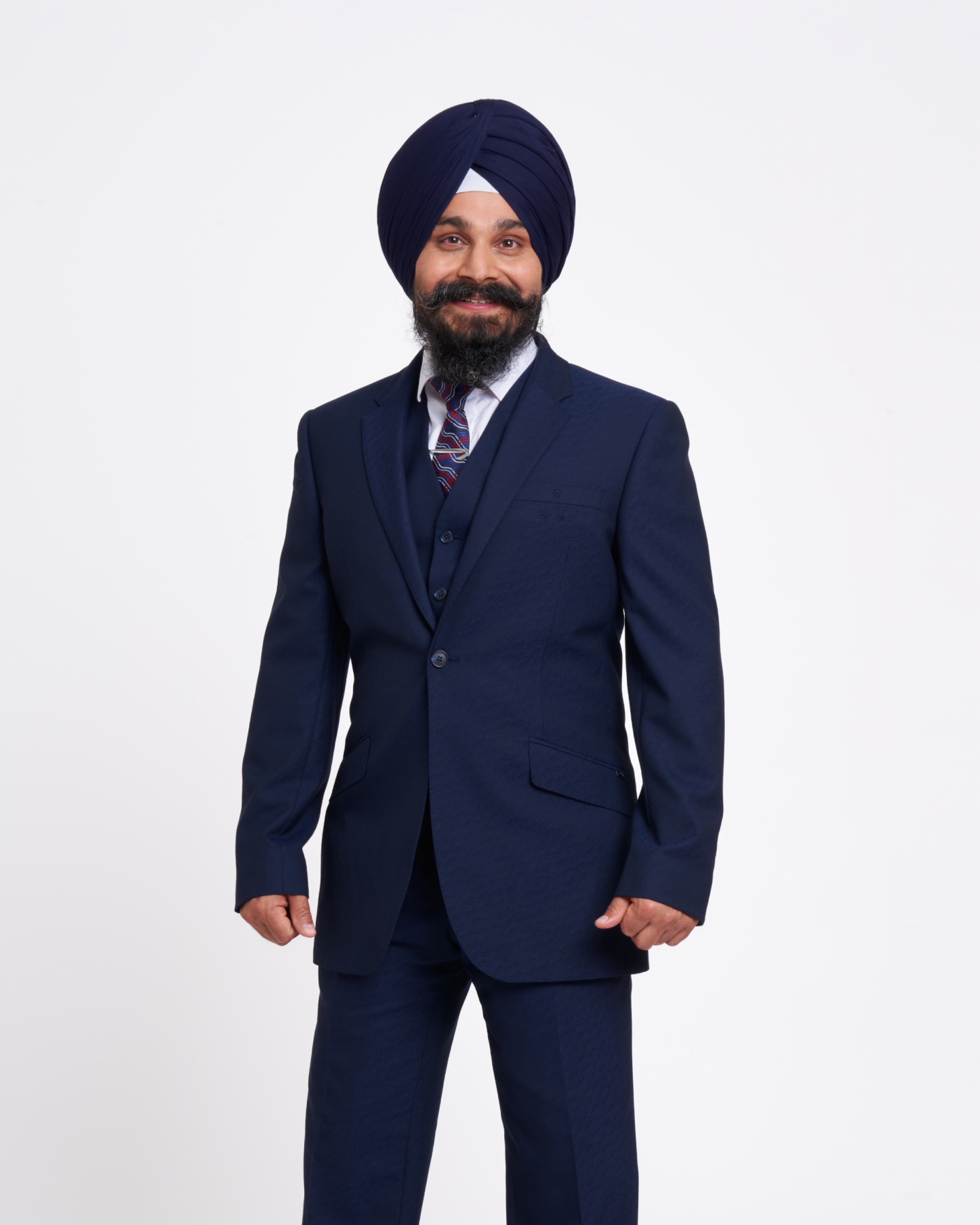 Smiling man wearing a navy blue three-piece suit and striped tie, with a dark turban, posing against a plain white background.