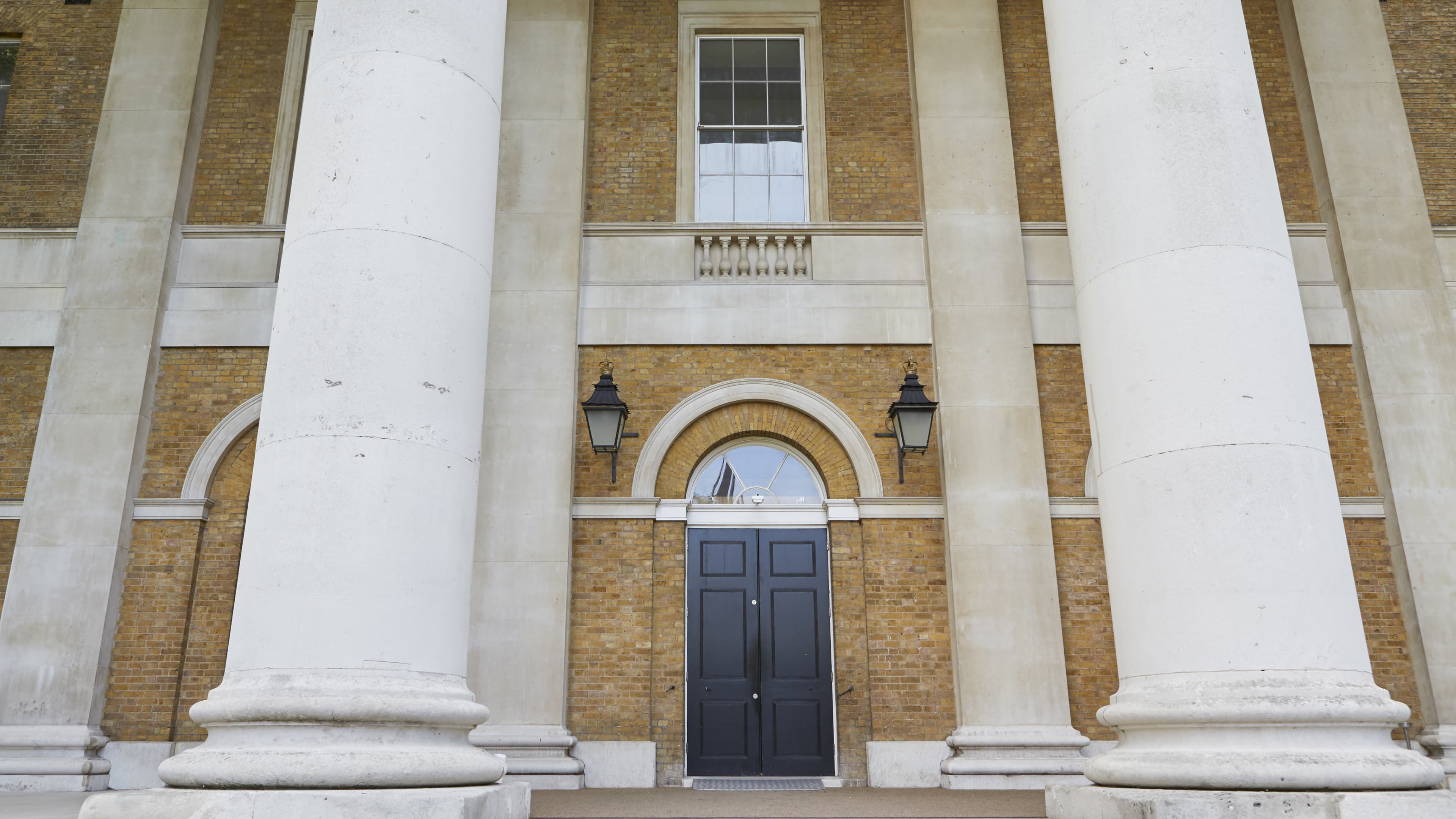 Tall white columns flank a large black door set in a brick wall, with two lanterns and a window above in a neoclassical building facade.