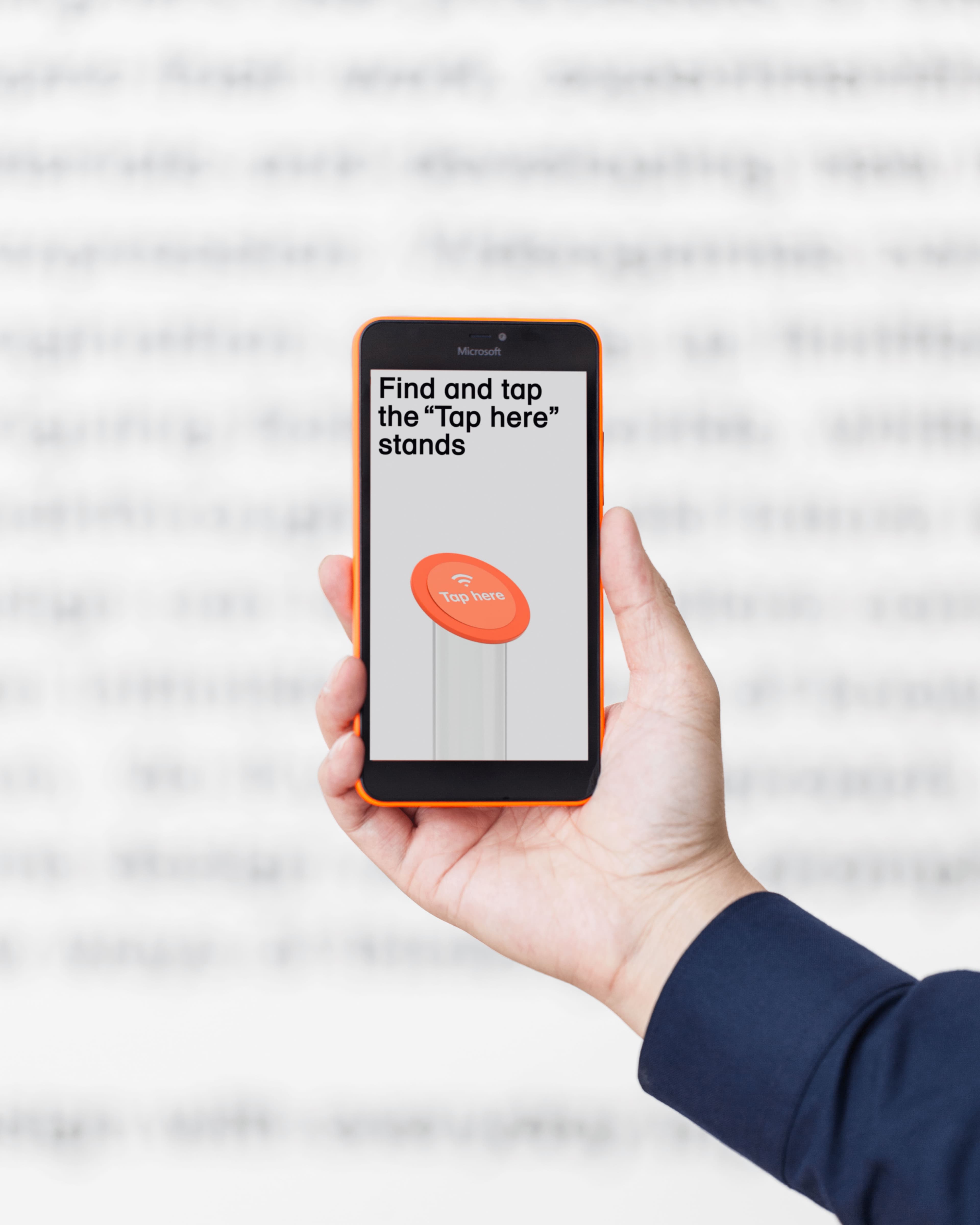 Hand holding an orange Microsoft Lumia smartphone displaying a screen instructing users to find and tap NFC "Tap here" stands, against a blurred text background.