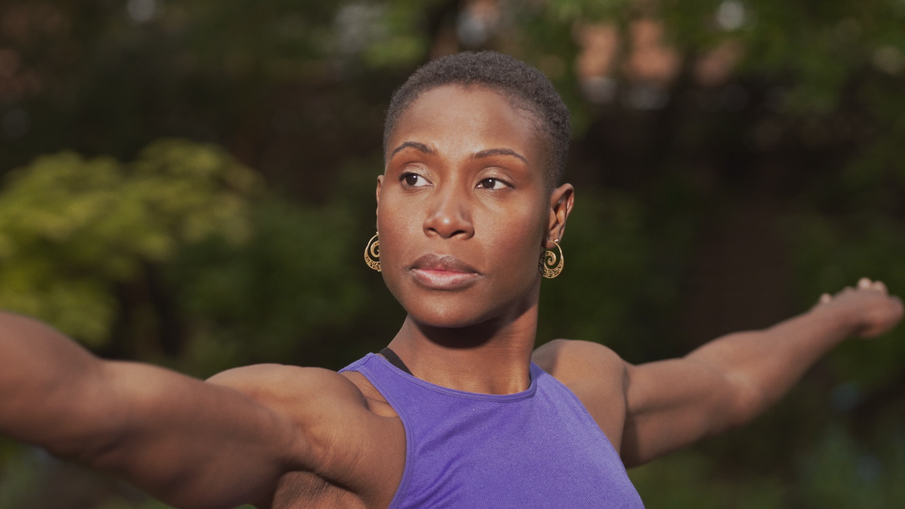 Black woman with short hair wearing a purple tank top and gold spiral earrings, arms extended in a yoga pose outdoors with greenery in the background.