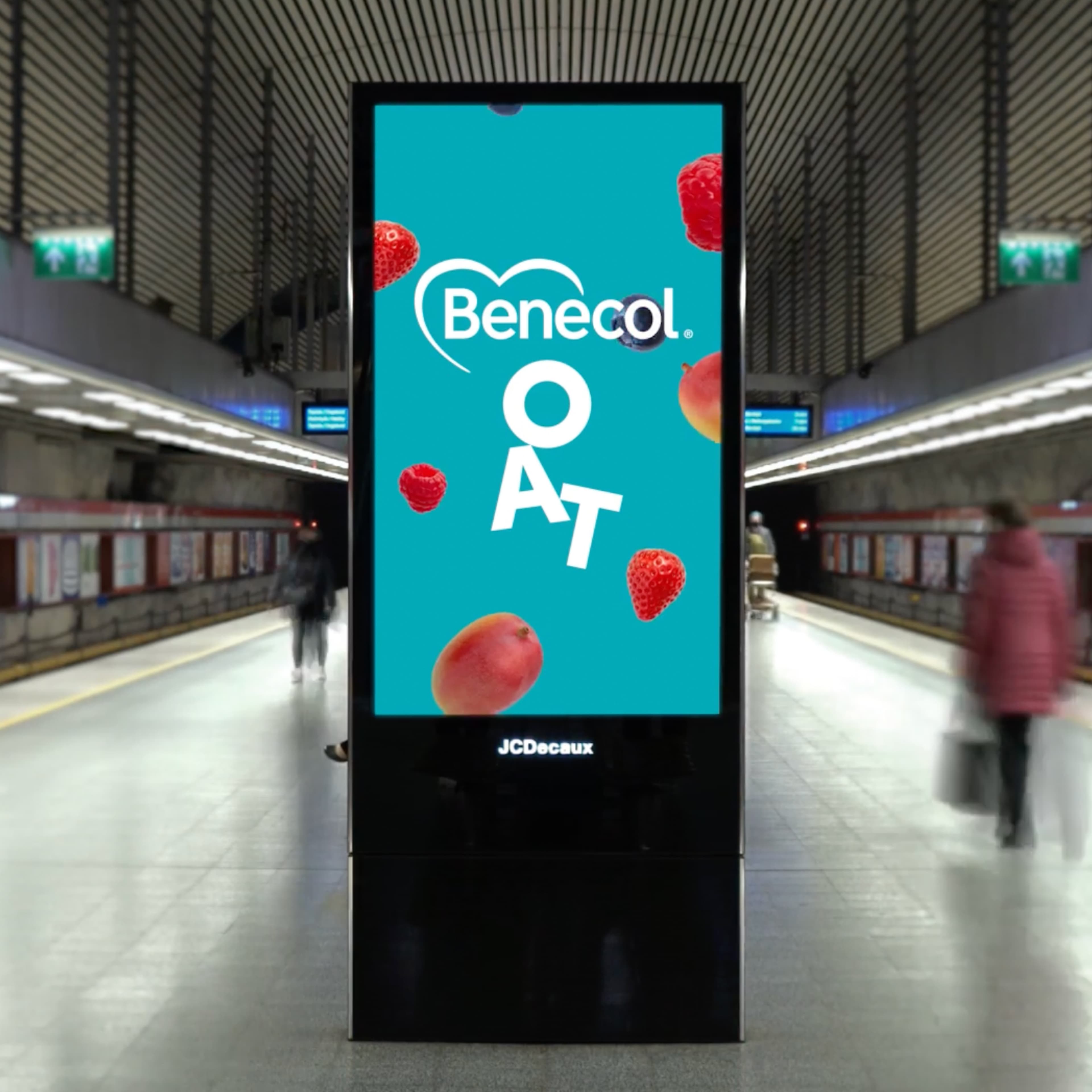 JCDecaux digital kiosk in a train station displaying a Benecol Oat advertisement with strawberries, raspberries, blueberries, and mangoes on a teal background, with commuters passing by.