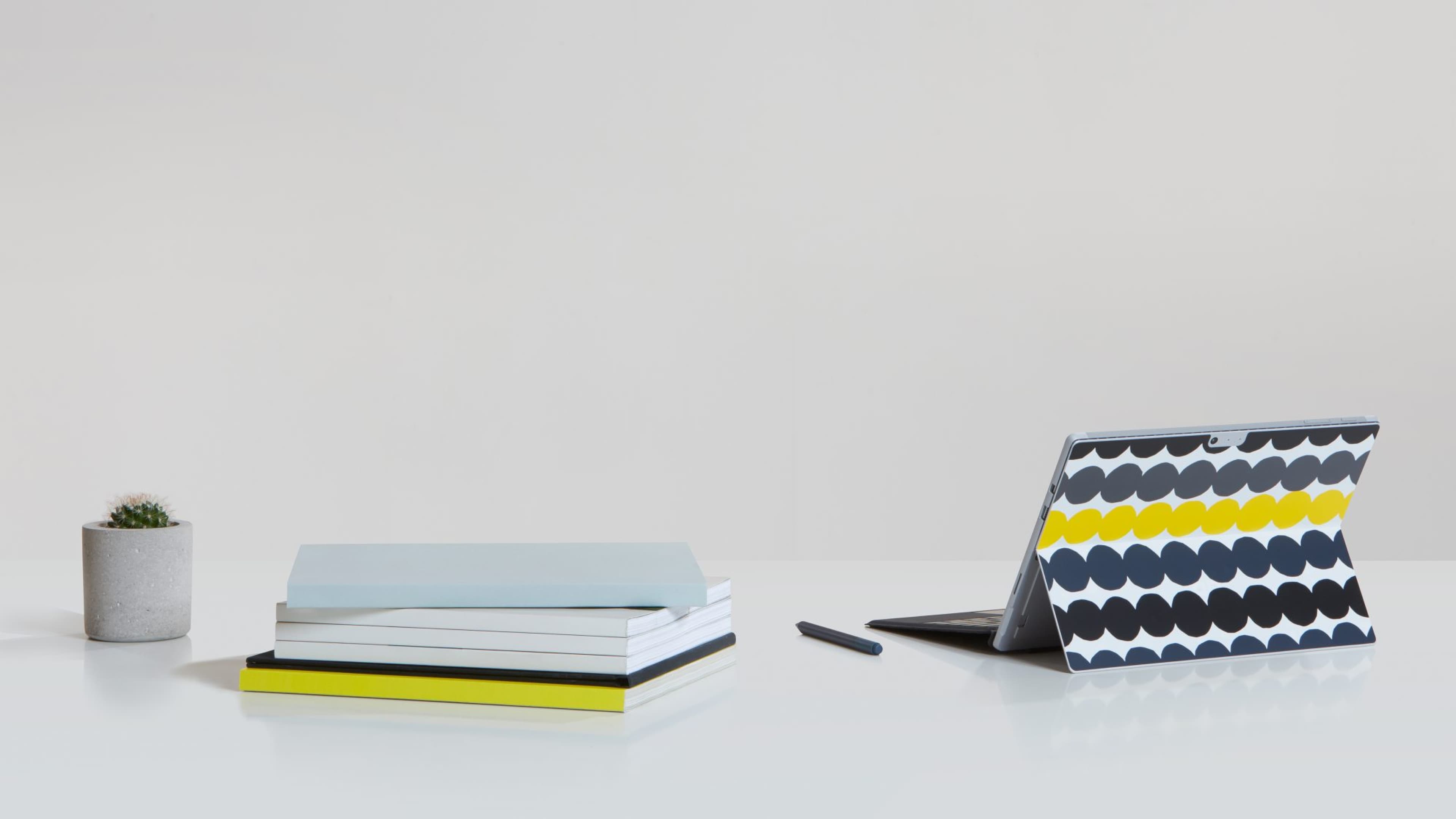 Microsoft Surface tablet with Marimekko dotted pattern cover in navy, yellow, and white, displayed on a white desk alongside a Surface Pen, stacked notebooks, and a small cactus in a concrete pot.