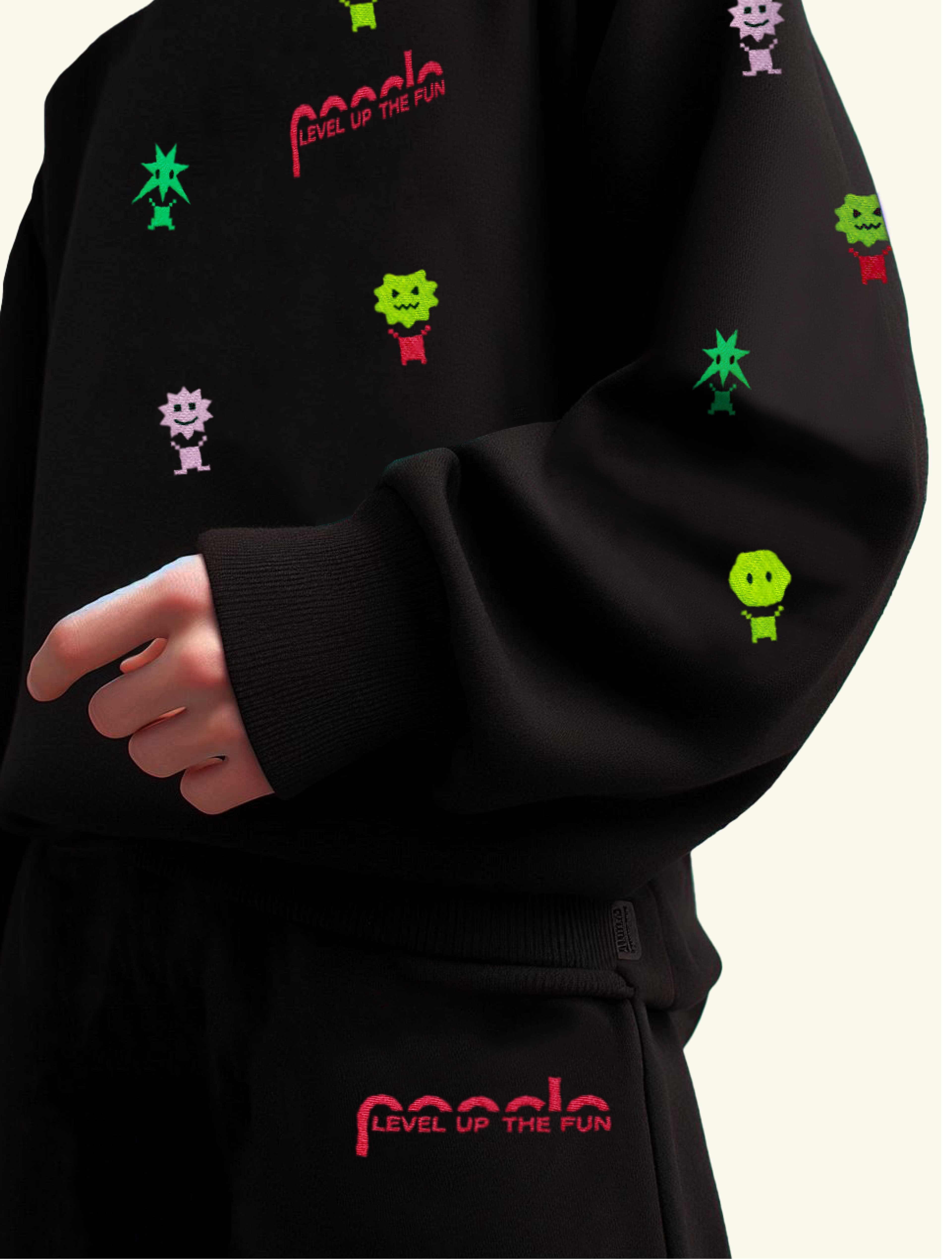 Close-up of a black matching sweatshirt and pants set with embroidered colorful pixel monster characters and red "Poncle – Level Up the Fun" text on the chest and lower hem.