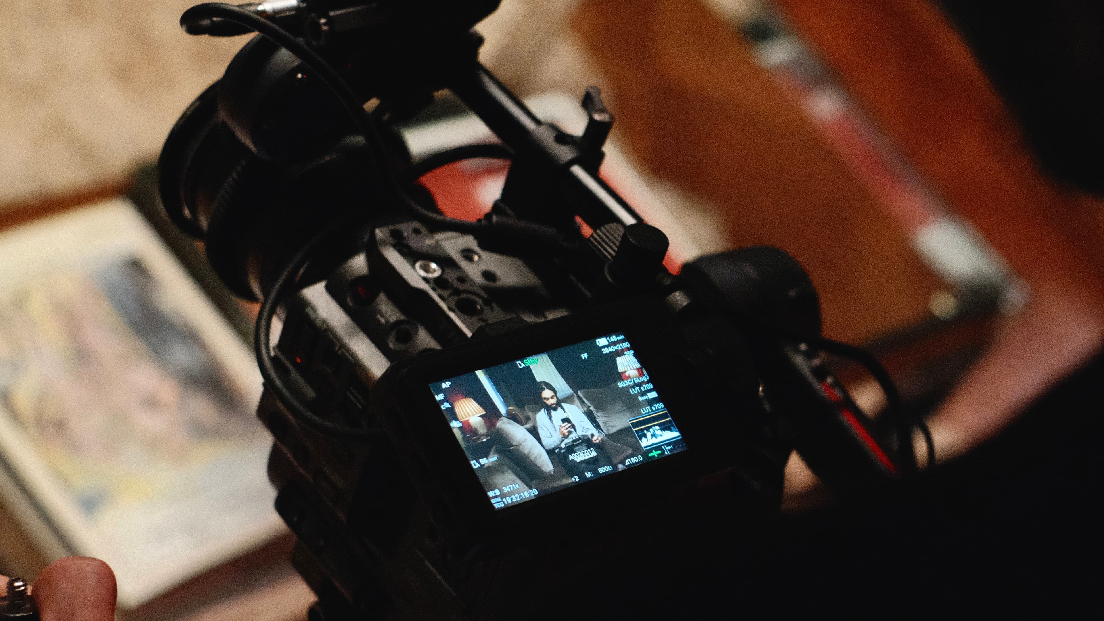Close-up of a video camera screen showing a person sitting on a couch with a lamp beside them. Camera controls visible in the foreground.