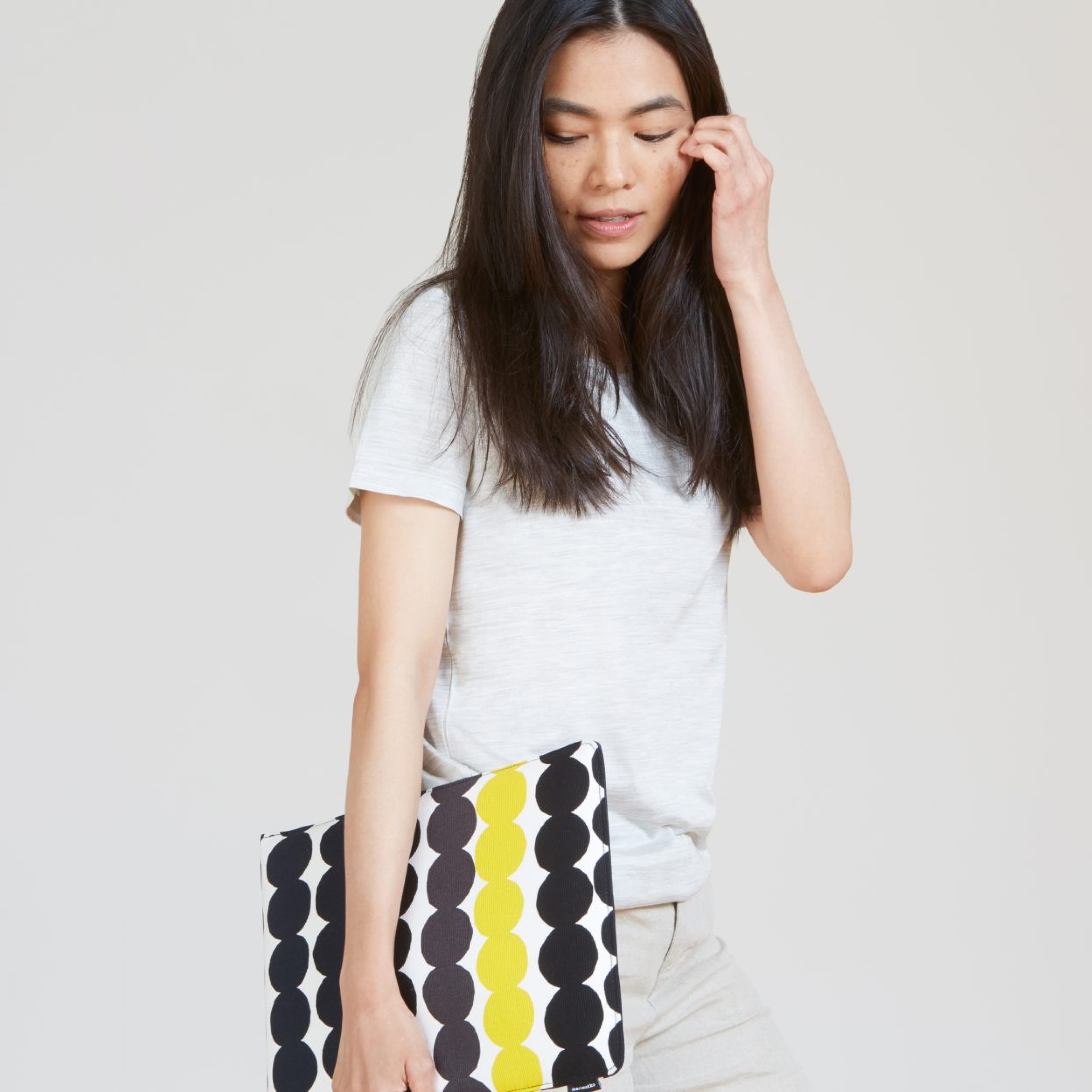 Woman with long dark hair wearing a white t-shirt, holding a Marimekko clutch pouch with black and yellow circular dot pattern, against a neutral gray background.